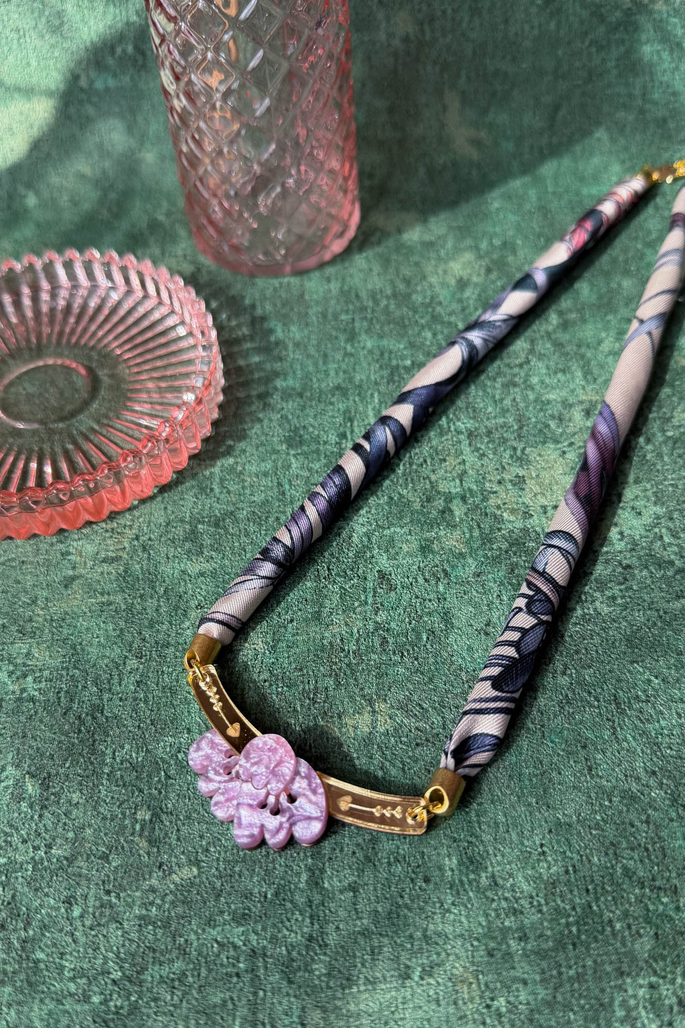 A handmade necklace featuring a patterned fabric and pink carved pendant with gold accents is displayed on a textured green surface, beside two pink glass dishes.