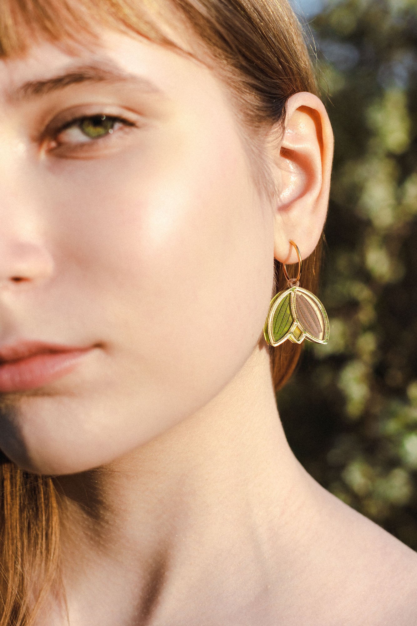 Close-up of a young woman’s face in natural light, focusing on her left eye, cheek, and ear. She wears lightweight handcrafted earrings shaped like gold and green leaves, with a blurred outdoor background.