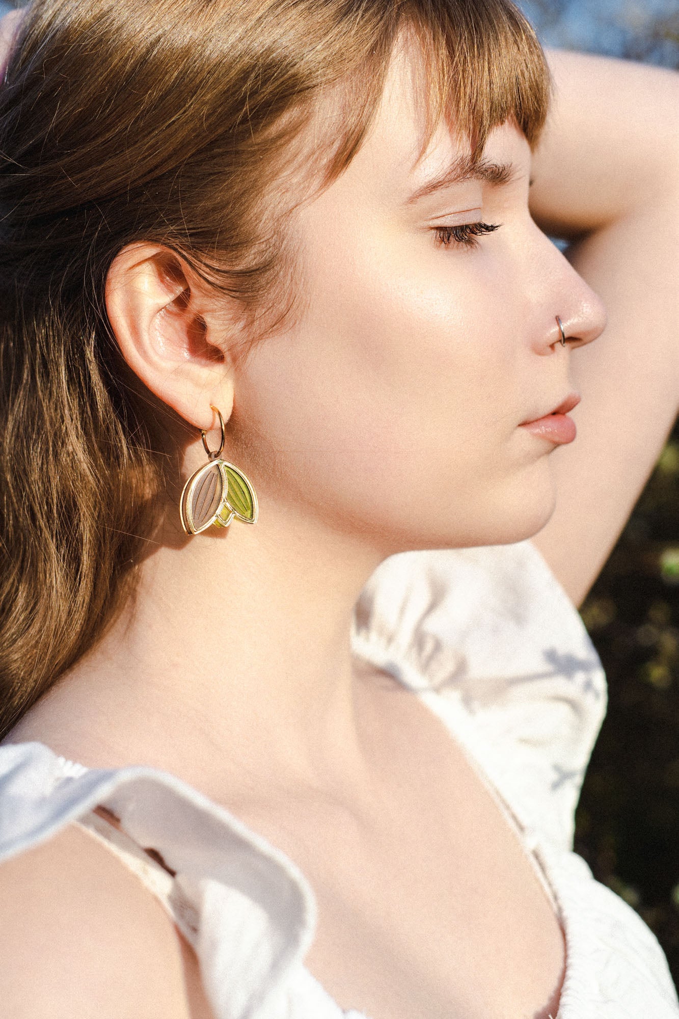 A young woman with light brown hair and fair skin stands outdoors, eyes closed, wearing a nose ring and lightweight handcrafted earrings. She is dressed in a white top with ruffled sleeves, one arm raised behind her head.