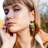 A young woman with light brown hair and bangs gazes calmly at the camera. She wears a tan fleece jacket and green and gold Art Nouveau earrings with a pearl drop. Her hand rests gently on her face, trees blurred in the background.