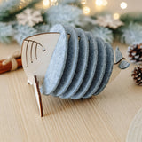 A set of cute coasters held in a wooden holder shaped like a whale, placed on a light wooden table with pine cones, fir branches, and warm fairy lights in the background—a charming touch for your home decor.