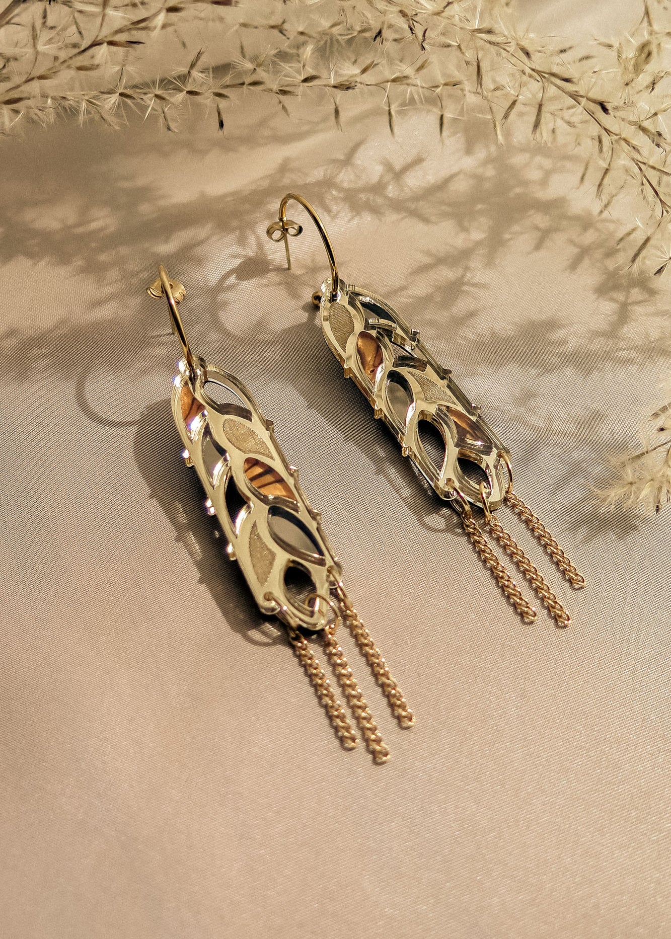 A pair of intricate gold and silver Wheat Statement Earrings with leaf-like cutouts and dangling chain accents, displayed on light beige fabric with delicate dried plants in the background for a standout style.