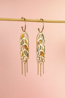 A pair of Wheat Statement Earrings with cut-out leaf patterns and dangling chain accents hang from a horizontal rod against a soft pink background, showcasing standout style.